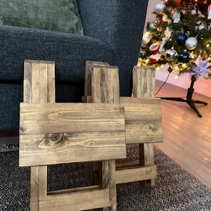 Op de afbeelding: Twee kleine, rustieke houten tafels met een natuurlijke afwerking. De tafels zijn geconstrueerd met een kruisvormig ontwerp en staan op een patroonkleed. Een donkergrijze fauteuil en een versierde kerstboom staan op de achtergrond.