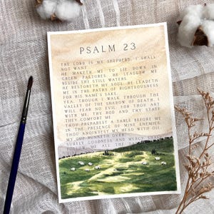 May include: Watercolor print of Psalm 23 with a landscape scene of sheep grazing in a green pasture. The text is in black, with a watercolor wash background in shades of brown and beige. A paintbrush and cotton bolls are in the background.