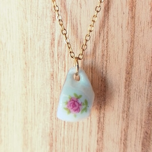 May include: A gold chain necklace with a porcelain teacup pendant featuring a pink rose design.