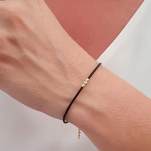 Puede incluir: Una pulsera de cordón negro con tres pequeñas cuentas doradas. La pulsera es sencilla y elegante, con un delicado cierre de cadena dorada. La pulsera se lleva en la muñeca.
