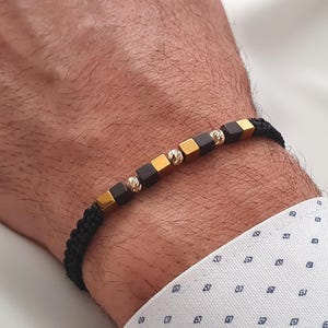 Könnte beinhalten: Schwarz geflochtenes Armband mit goldenen und dunkelgrauen Würfelperlen sowie strukturierten goldenen Perlen. Das Armband befindet sich an einem Handgelenk, wodurch das Design hervorgehoben wird. Ein stilvolles Accessoire.