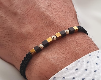 Puede incluir: Pulsera trenzada negra con cuentas cúbicas doradas y gris oscuro, y cuentas doradas texturizadas. La pulsera se muestra en una muñeca, mostrando el diseño. Un accesorio elegante.