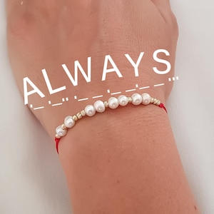 Peut inclure: Un bracelet délicat avec des perles blanches et des perles dorées montées sur un cordon rouge. Le mot "ALWAYS" est affiché au-dessus du bracelet en blanc, avec une représentation en code Morse en dessous.