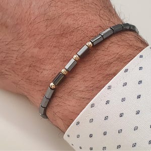 May include: A dark gray beaded bracelet with gold-colored accents. The bracelet is made of rectangular beads and small, round gold beads. The bracelet is worn on a wrist, with a white shirt with a blue pattern in the background.