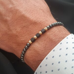 May include: A beaded bracelet featuring small, dark gray beads interspersed with gold-colored, textured beads and dark brown square beads. The bracelet is worn on a wrist, showcasing the contrast of colors and textures.
