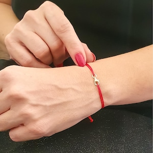 May include: A red string bracelet with a gold clasp. The bracelet is tied around a person's wrist.