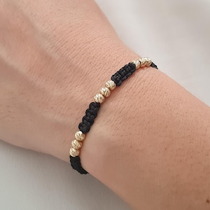 May include: A black string bracelet with gold beads. The beads are round and have a textured surface.