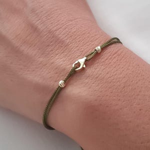 May include: Olive green cord bracelet with gold-coloured metal accents. The bracelet features two small gold beads and a clasp. The bracelet is designed to be worn on the wrist.