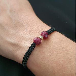 May include: A black braided bracelet with two red gemstone beads. The beads are faceted and have a gold accent.