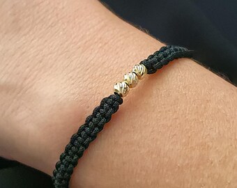 May include: Black braided bracelet with three gold beads. The bracelet is worn on a wrist, showcasing the contrast between the dark cord and the shiny gold accents. A simple, elegant piece of jewelry.