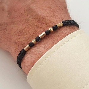 Könnte beinhalten: Schwarz geflochtenes Armband mit goldfarbenen rechteckigen und kugelförmigen Perlen. Das Armband wird am Handgelenk getragen, mit einem cremefarbenen Hemdsärmel im Hintergrund. Ein stilvolles Accessoire.