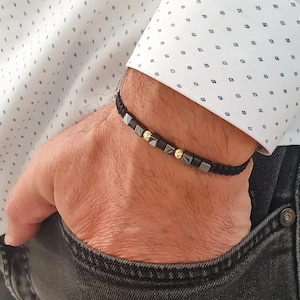 May include: Black braided bracelet featuring square and round beads in silver and gold tones. The bracelet is worn on a wrist, showcasing the jewelry's design and texture. The bracelet is a stylish accessory.