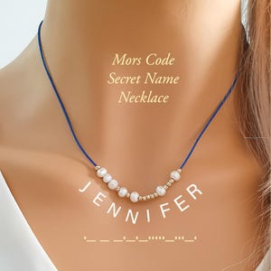 May include: A blue cord necklace with white pearls and gold beads. The word "JENNIFER" is spelled out below the necklace, with the Morse code equivalent displayed underneath. The text "Mors Code Secret Name Necklace" is also visible.