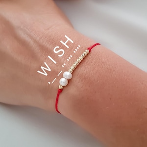 Puede incluir: Pulsera de hilo rojo con cuentas doradas y dos perlas blancas. La palabra "WISH" está impresa en blanco en el lado izquierdo de la imagen. La pulsera está atada a la muñeca.