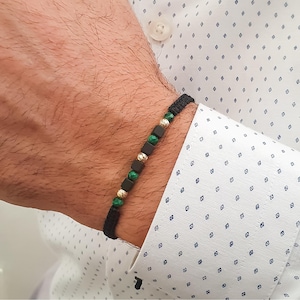 May include: Black woven bracelet with gold-colored beads, dark green beads, and dark square beads. The bracelet is worn on a wrist, and the background includes a white shirt with a blue pattern.