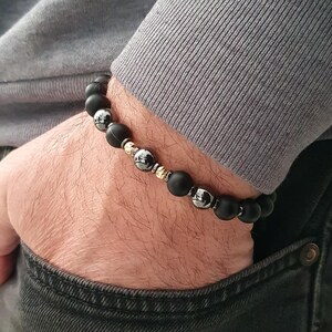 May include: A black beaded bracelet with gold and silver accents. The bracelet is made of matte black beads, interspersed with gold-colored beads and silver-colored beads. The bracelet is worn on a wrist.