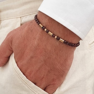 Può includere: Un braccialetto di perline con piccole sfere rosso scuro alternate a distanziatori cilindrici color oro e minuscoli accenti sferici dorati, che creano un motivo ripetitivo lungo l'intera lunghezza del gioiello.