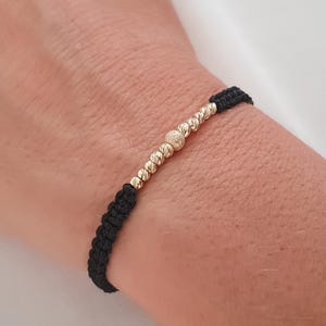Könnte beinhalten: Ein schwarzes gewebtes Armband mit goldfarbenen Perlen. Das Armband hat eine Reihe kleiner, runder Perlen und zwei größere, strukturierte Perlen. Das Armband wird am Handgelenk getragen.