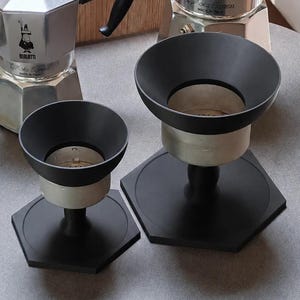 Könnte beinhalten: Zwei schwarze, trichterförmige Kaffeedosierringe mit sechseckigen Sockeln, platziert vor zwei silbernen Bialetti Moka Express Espressokochern. Die Dosierringe verfügen über eine breite, trichterförmige Öffnung und ein Standfuß-Design.