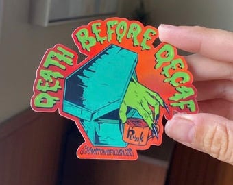 Puede incluir: Una pegatina troquelada que muestra un ataúd de color verde azulado con una mano verde saliendo de él, sosteniendo una taza naranja con la palabra "Punk". El texto "DEATH BEFORE DECAF" aparece en una fuente verde que parece gotear.