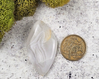 May include: A polished, teardrop-shaped gemstone with a translucent, layered appearance in shades of white and gray. A gold-colored coin is next to the gemstone. The gemstone is displayed on a textured, light gray surface.
