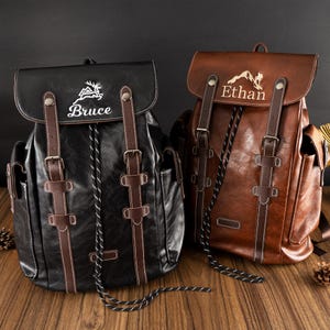 May include: Two leather backpacks, one black and one brown, with buckle fastenings and white stitching. The black backpack has the name "Bruce" embroidered on the flap, and the brown backpack has the name "Ethan". Both have a mountain design.