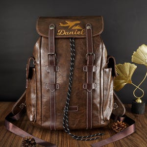 May include: A dark brown faux leather backpack featuring two vertical straps with metal buckles, a drawstring closure, and side pockets. The top flap is embroidered with a gold aeroplane icon and the name Daniel in a cursive script font.