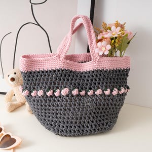 Könnte beinhalten: Handgefertigte Häkeltasche in Grau und Rosa. Die Tasche hat einen rosa oberen Rand und Henkel, mit einer Reihe von erdbeerförmigen Verzierungen. Die Tasche ist mit rosa Blumen gefüllt.
