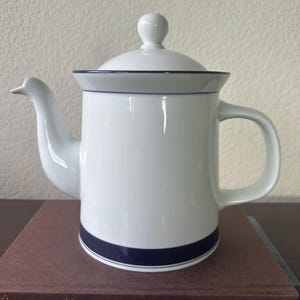 May include: White ceramic teapot with a blue trim and a lid. The teapot has a handle and a spout. The teapot is sitting on a brown book with the title "Happy Alchemy" on the cover.