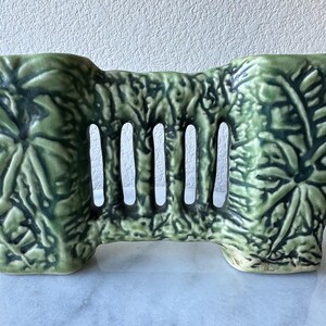 May include: A green ceramic planter with a rectangular shape and a series of vertical openings. The planter is decorated with a leaf pattern. The planter is approximately 8 inches wide and 5 inches tall.