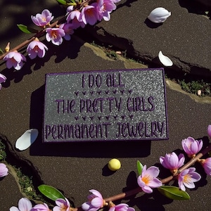 May include: A rectangular sign with the text "I DO ALL THE PRETTY GIRLS PERMANENT JEWELRY" in purple lettering on a silver glitter background. The sign is surrounded by pink flowers and petals.