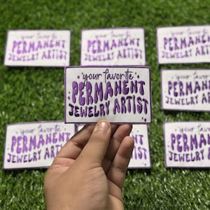May include: Rectangular white patches with purple embroidered text that reads "your favorite PERMANENT JEWELRY ARTIST." Several patches are visible, with one held by a hand, set against a green background.