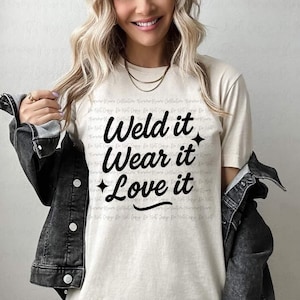 Op de afbeelding: Crèmekleurig T-shirt met de tekst "Weld it, Wear it, Love it" in zwart script. Het shirt heeft een ronde hals en korte mouwen. Een zwarte spijkerjas is over de schouders gedrapeerd.