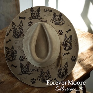 May include: Tan fedora-style hat with a wide brim, decorated with German Shepherd dog portraits, paw prints, and bone graphics. The word "LOVE" is printed on the brim. The hat is on a wooden surface.