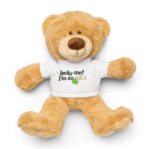 Puede incluir: Un oso de peluche marrón claro con una camiseta blanca. La camiseta tiene el texto "lucky me! I'm an AKA" con un trébol verde y texto rosa y verde. El oso tiene una nariz marrón y ojos negros.