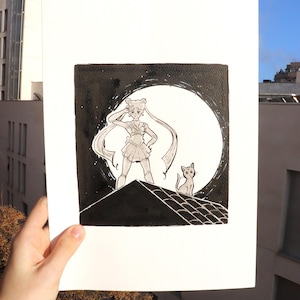 Può includere: Illustrazione in bianco e nero di un personaggio in uniforme da marinaio in piedi su un tetto di fronte a una grande luna. Anche un gatto è sul tetto. L'opera d'arte è tenuta contro uno sfondo di cielo blu.