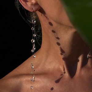 Peut inclure: Une longue boucle d'oreille pendante composée d'une série de perles de verre transparentes, sphériques et en forme de goutte, suspendues à de fins filaments métalliques délicats. Le design crée un effet de cascade avec des perles variées.