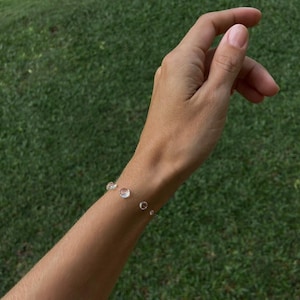 May include: A delicate bracelet featuring clear, round gemstones of varying sizes. The bracelet is strung on a thin, silver-colored chain. The background is a blurred view of green grass.