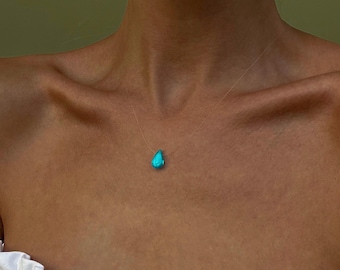 May include: A delicate necklace featuring a teardrop-shaped turquoise pendant. The pendant is suspended on a nearly invisible clear cord, creating a floating effect. The necklace is worn against a skin tone background.