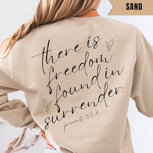May include: Sand-colored sweatshirt with the text "there is freedom found in surrender" in black script. The text is accompanied by two small heart symbols. The scripture reference "proverbs 3:5-6" is below the main text.