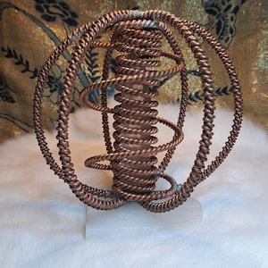 May include: An abstract sculpture in copper tones, with a spherical shape. The design incorporates numerous spiraling and circular components, forming an open, airy structure. It rests on a white rectangular base, set against a textured, golden backdrop.