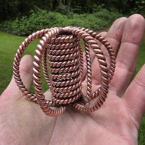May include: A copper wire sculpture featuring a central coil with multiple loops extending outwards. The sculpture is crafted from twisted copper wire and has a reddish-brown hue.