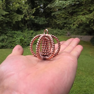 Könnte beinhalten: Ein kupferfarbener Anhänger mit einem einzigartigen, gewebten Design. Der Anhänger hat zwei äußere Ringe und einen inneren Kern, die alle aus verdrilltem Metall gefertigt sind. Das Schmuckstück wird in einer Hand vor dem Hintergrund grüner Blätter gehalten.