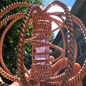 May include: A copper-coloured wire-wrapped sculpture with a central, elongated, light purple crystal. The wire is intricately woven into a spherical cage-like structure. The background includes green foliage and a bright blue sky.