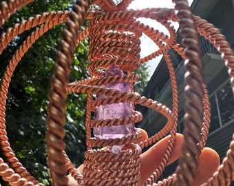 Könnte beinhalten: Eine kupferfarbene, drahtgewickelte Skulptur mit einem zentralen, länglichen, hellvioletten Kristall. Der Draht ist kunstvoll zu einer kugelförmigen, käfigartigen Struktur verwoben. Der Hintergrund umfasst grünes Laub und einen hellblauen Himmel.