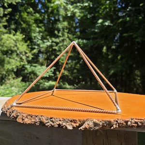 May include: A copper-toned pyramid structure with a square base and triangular apex. The pyramid is made from twisted metal rods and rests on a wooden slab with a natural edge. The background shows green trees and a clear blue sky.