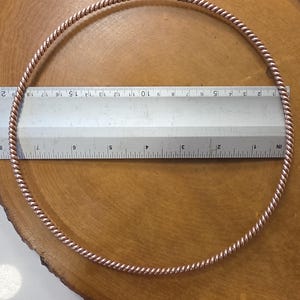 Op de afbeelding: Een koperkleurige, gedraaide metalen hoepel, ongeveer 20 cm in diameter, ligt op een houten oppervlak. Een liniaal is over de hoepel geplaatst voor meting. Het ontwerp van de hoepel heeft een spiraalpatroon en de algehele esthetiek is rustiek en handgemaakt.