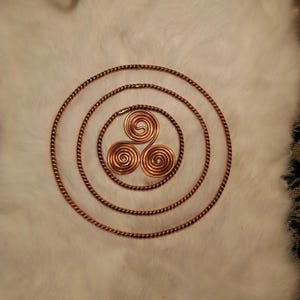 Puede incluir: Tres espirales de alambre de cobre, o triskelions, están dispuestas en un patrón triangular. Las espirales están rodeadas por tres círculos concéntricos de alambre de cobre.
