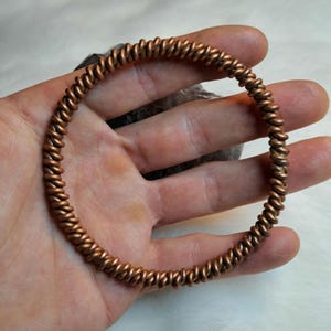 Op de afbeelding: Een koperen draad armband met een gedraaid ontwerp. De armband is gemaakt van een enkel stuk draad dat is gedraaid tot een spiraalvorm.