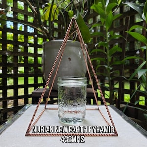 Op de afbeelding: Een koperkleurige piramidestructuur omringt een heldere glazen pot gevuld met water. De pot staat in het midden van de piramide, die op een wit oppervlak is geplaatst. De tekst onderaan luidt "NUBIAN NEW EARTH PYRAMID 432MHZ".